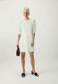 Vestido curto branco com detalhes de franjas texturizadas e mangas bufantes. Combinado com sandálias de salto pretas e uma pequena bolsa bordeaux.