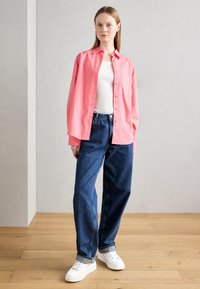 Light pink button-up shirt over a white t-shirt, paired with blue high-waisted jeans and white sneakers. Wood floor background.