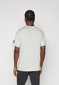 Ανοιχτό γκρι μπλουζάκι φτιαγμένο από μαλακό ύφασμα, με στρογγυλή λαιμόκοψη και κοντά μανίκια, με μπλε έμβλημα στο αριστερό μανίκι.