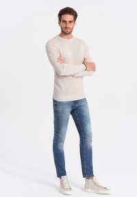 Pull en maille beige clair, jean slim bleu, et baskets montantes beige clair. Le modèle se tient les bras croisés devant un fond uni.