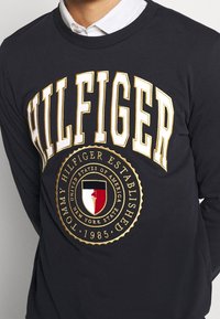 Laivastonsininen puuvillapaita, jossa on rohkea valkoinen ja kultainen "HILFIGER"-teksti. Sisältää pyöreän logon, jossa on kynttilän design. Pyöreä pääntie ja pitkät hihat.