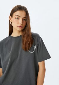 Camiseta de color gris carbón en tejido suave con un ajuste relajado. Presenta un diseño gráfico en blanco en el lado izquierdo del pecho, que representa una forma de corazón.
