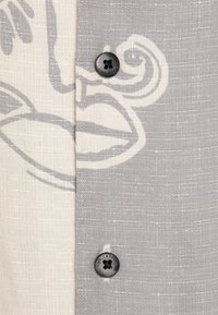 Camisa de botones que presenta un diseño dividido de tela gris claro y blanca con un estampado negro. Dos botones, uno negro con un detalle floral.