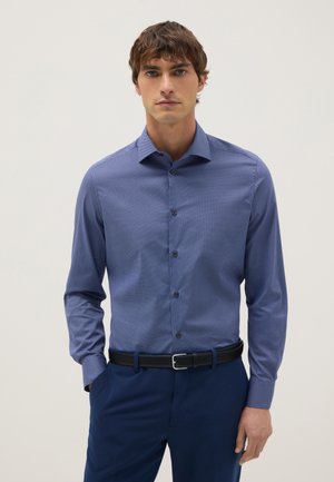 Hombre vestido con una camisa azul ajustada de textura acanalada con botones y mangas largas, metida dentro de pantalones azul marino con cinturón negro, de pie contra un fondo liso.