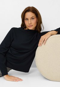 Zwart, top met lange mouwen, een hoge hals en een losse pasvorm. Een rond kussen met textuur in beige is gedeeltelijk zichtbaar naast de hand van het model.