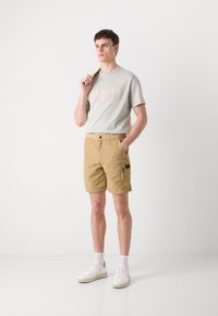 Tricou gri deschis cu text roz în relief "BARBOUR INTERNATIONAL", asociat cu pantaloni scurți cargo bej și adidași albi.