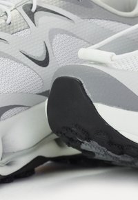 Încălțăminte sport gri și albă, cu un material superior din plasă texturată, design curbat și talpă din cauciuc cu modele de aderență. Dispune de șireturi și accente cu logo.