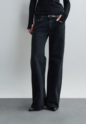 Osoba ubrana w czarne dżinsy z wysokim stanem i szerokimi nogawkami, czarny pasek, czarne buty w szpic oraz dopasowany czarny top z długim rękawem.