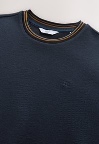 Felpa blu navy con scollo rotondo in tessuto testurizzato con polsini a coste. Presenta un bordo del collo a strisce dorate e nere e un piccolo emblema a forma di corona.