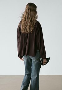 Weinrotes, übergroßes Hemd mit abgerundetem Saum, kombiniert mit hellblauen Jeans. Das Hemd hat lange Ärmel und eine lockere Passform.