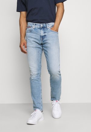 Mann trägt hellblaue Slim-Fit-Jeans, navyblaues Hemd und weiße Sneaker, steht mit einer Hand in der Tasche vor einem einfarbigen Hintergrund.