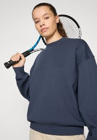 Marineblaue Sweatshirt mit Rundhalsausschnitt und überschnittenen Schultern, mit gesticktem Logo auf der Brust. Das Model hält einen Tennisschläger.