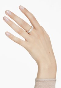 En hand med manikyrerade naglar visar en guldring prydd med flera klara stenar. Bakgrunden är enkel vit.