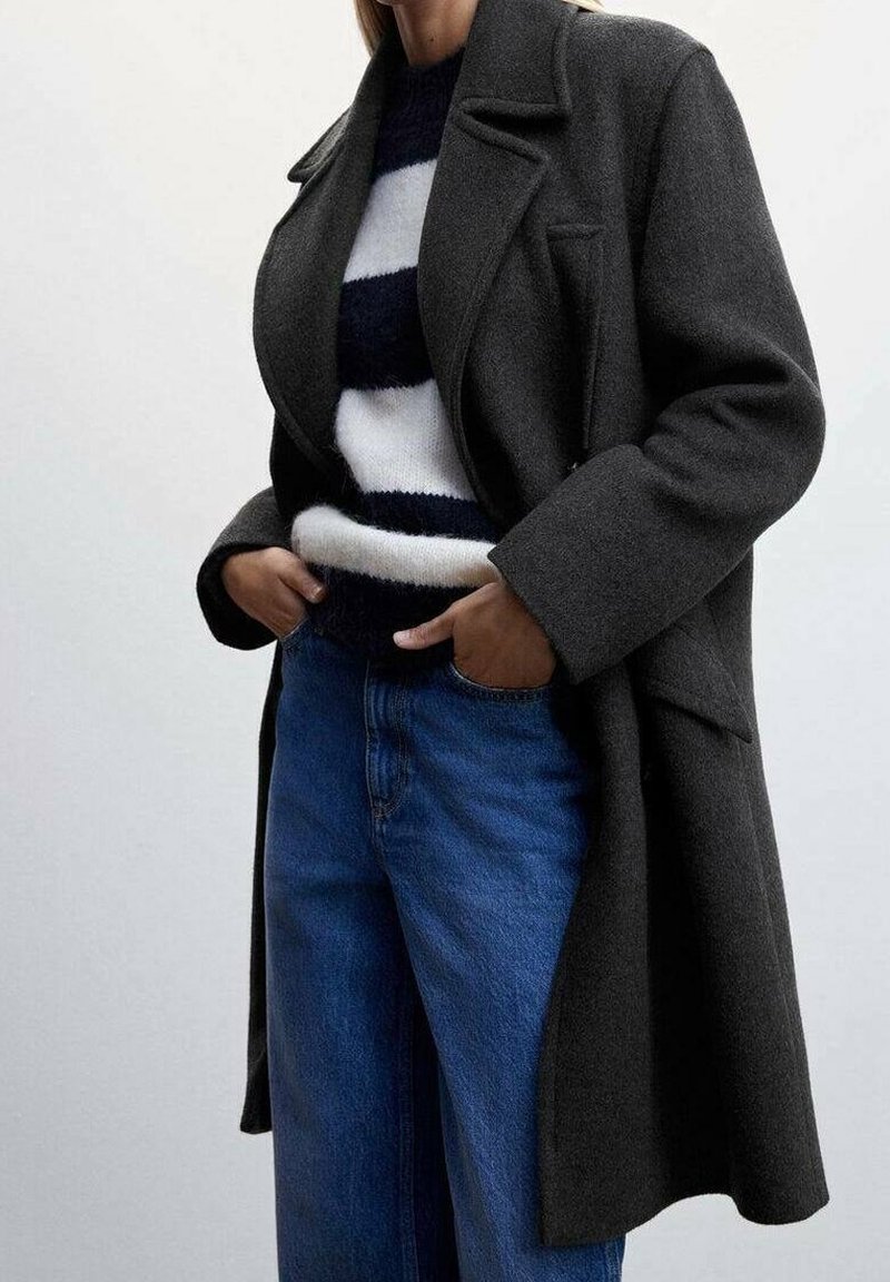 Persoon in een donkergrijze lange jas over een zwart-wit gestreepte trui en blauwe spijkerbroek, met handen in de zakken, staande tegen een effen achtergrond.