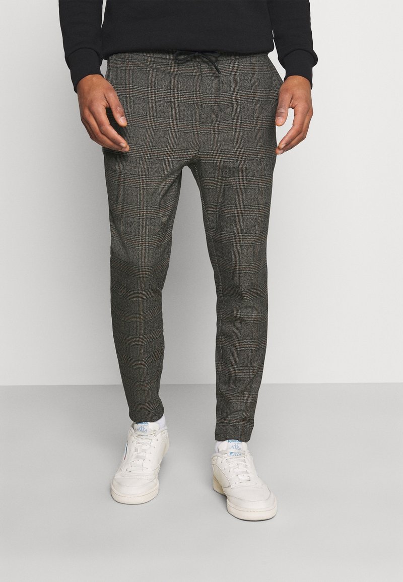 Pantalones de cuadros grises con cintura ajustable, corte tapered y tela texturada, combinados con zapatillas blancas y calcetines blancos hasta el tobillo.