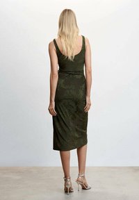 Robe midi sans manches vert olive, avec une silhouette ajustée et de subtils motifs végétaux. Portée avec des talons argentés à lanières, mettant en valeur une texture lisse.