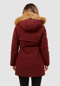 Parka marrone con bordo in pelliccia sintetica, dotata di vita sfiancata, maniche lunghe e una superficie liscia. Presenta un dettaglio di ventilazione sul retro.