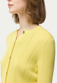 Gul ribbade cardigan med v-halsringning, försedd med en dekorativ silverknapp vid kragen. Mjuk textur och figurnära design.