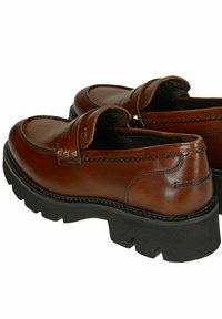 Mocassini in pelle marrone con suole in gomma nera spessa, cuciture decorative e borchie metalliche triangolari sul dettaglio della strap.