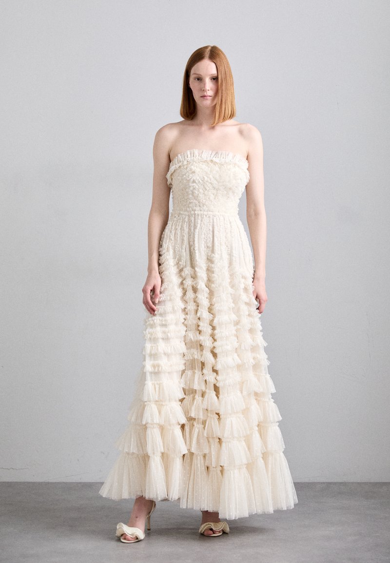 Robe ivoire sans bretelles mettant en valeur un tissu texturé, des volants superposés et une silhouette fluide ; associée à des sandales à talons crème.