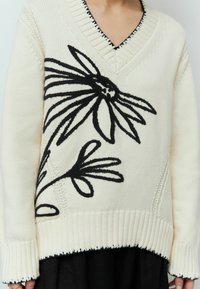 Pull en tricot crème avec un motif floral noir, décolleté en V, parements côtelés, et surpiqûres contrastées le long de l'ourlet et de l'encolure.