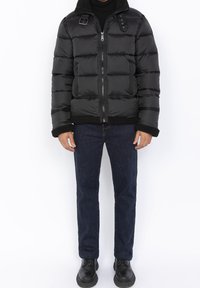 Czarna kurtka puchowa z zamkiem błyskawicznym z przodu, dużymi pikowanymi panelami, kołnierzem wykończonym futrem oraz kieszeniami po bokach, zestawiona z ciemnymi dżinsami i czarnymi butami.