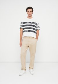 Λευκό μπλουζάκι με μαύρες οριζόντιες ρίγες και το λογότυπο "GUESS". Μπεζ παντελόνι με ίσια γραμμή. Το μοντέλο φοράει λευκά αθλητικά παπούτσια, στέκεται σε ουδέτερο φόντο.