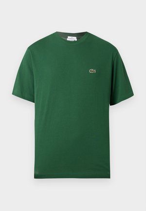 Homme et femme debout côte à côte portant des T-shirts Lacoste verts assortis avec le logo, la femme en jupe en jean bleu et baskets, l'homme en jean bleu et chaussures blanches.
