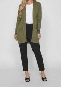 Olivgrön kofta med öppen fram, långa ärmar och sidofickor, parat med svarta byxor och svarta öppna klackar.