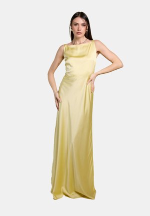 Donna in posa con un abito in raso giallo pallido lungo fino a terra, senza maniche, con scollatura quadrata, su uno sfondo semplice.