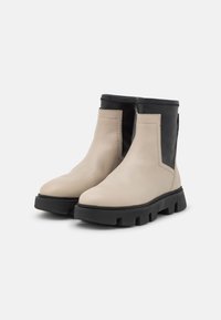 Botas de tobillo beige y negras con un acabado de cuero suave, paneles laterales elásticos y suelas de goma negra resistentes. Punta cuadrada y diseño robusto.