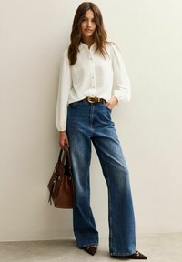 Femme portant un chemisier blanc, un jean large bleu, une ceinture marron, tenant un sac à main marron, se tenant contre un mur beige.