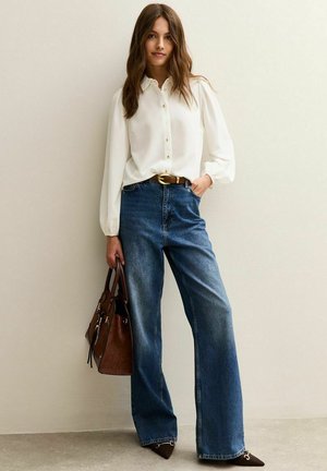 Woman wearing a white blouse, blue wide-leg jeans, brown belt, holding a brown handbag, standing against a beige wall.
