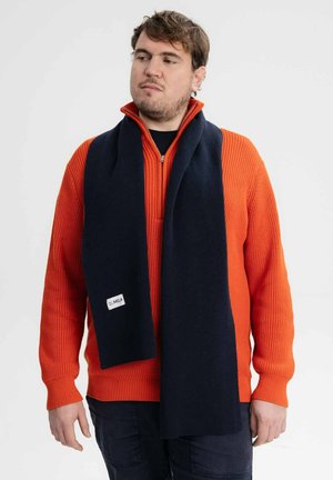 Navy sjaal met een gladde textuur om de nek gedrapeerd, voorzien van een klein wit label, gecombineerd met een gestructureerd feloranje sweater.