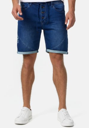 Dunkelblaue Denim-Shorts mit einem verblassten Look, ausgestattet mit einem Knopfverschluss, zwei vorderen Taschen und umgeschlagenen Säumen. Kombiniert mit weißen Sneakers.