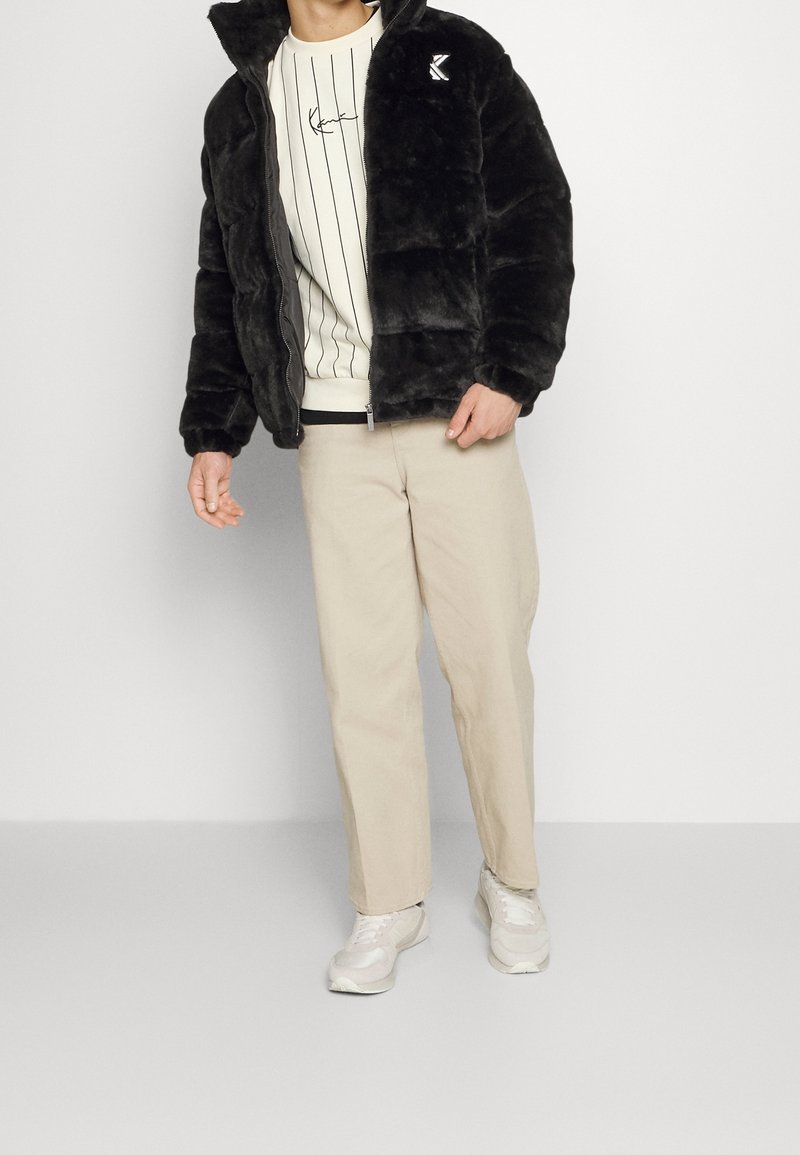 Homme portant une veste en fausse fourrure noire par-dessus un sweat-shirt à rayures blanches, pantalon beige et baskets blanches, debout devant un fond blanc uni.
