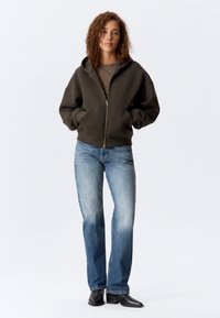 Dunkelgrüne Zip-Hoodie mit großen Taschen, getragen über einem braunen Shirt, kombiniert mit hellblauen Jeans und schwarzen Stiefeletten. Glatte Textur.