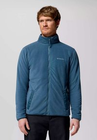 Giacca in pile di colore teal con colletto alto, chiusura con zip completa e tasche laterali. Presenta una texture liscia e un logo discreto sul petto.