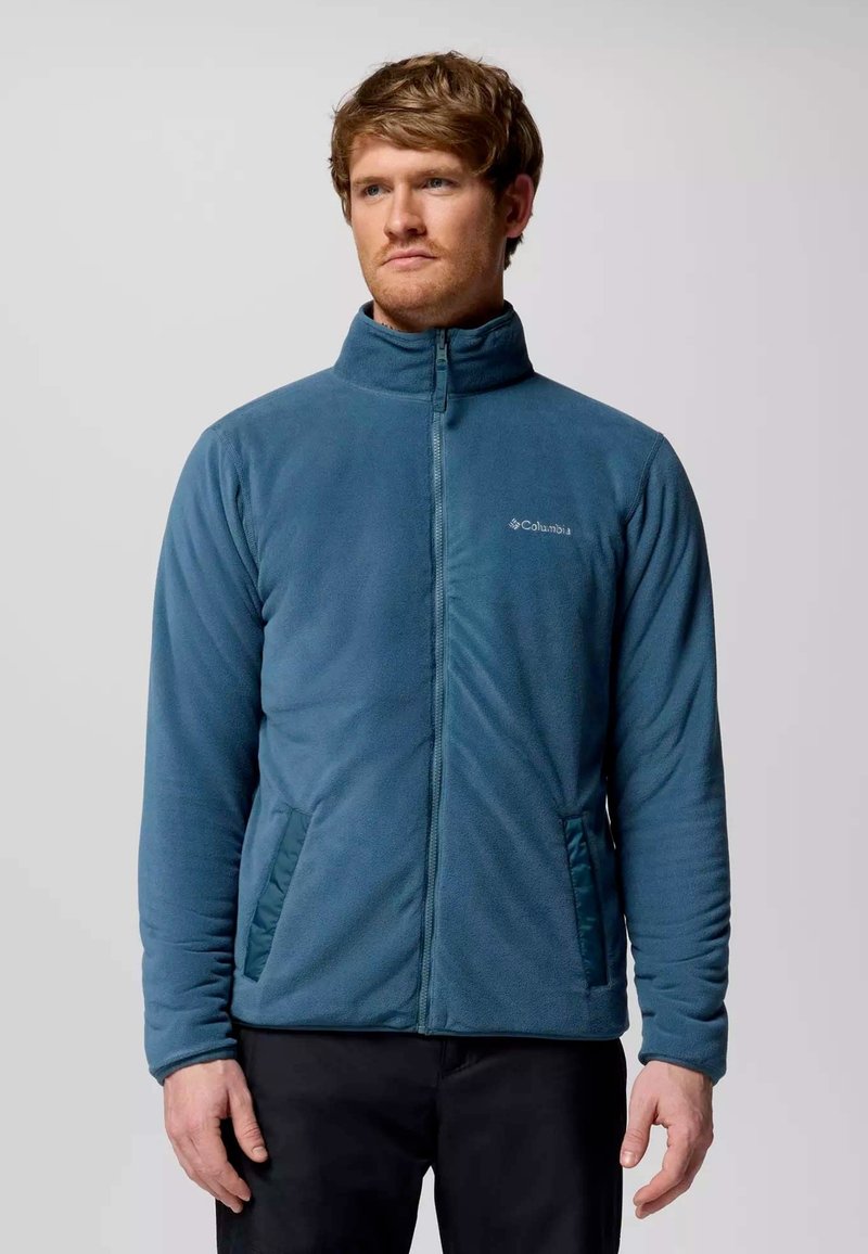 Giacca in pile di colore teal con colletto alto, chiusura con zip completa e tasche laterali. Presenta una texture liscia e un logo discreto sul petto.