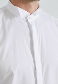 Chemise blanche avec un col montant, texture lisse et fermeture à boutons sur le devant avec une patte dissimulée pour un look épuré.