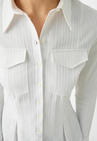 White fitted ribbed shirt with vertical stripes, buttoned front, collar, and flap chest pockets on a person.