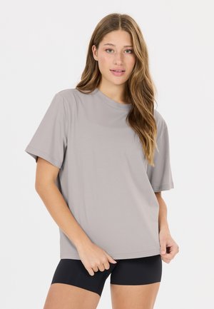 Ung kvinde med langt hår iført en løs, lys grå t-shirt og sorte shorts, står foran en ensfarvet hvid baggrund.
