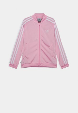 Różowa kurtka sportowa Adidas z białymi trzema paskami na rękawach, zamkiem błyskawicznym z przodu, dwoma bocznymi kieszeniami i logo Adidas trefoil na piersi.