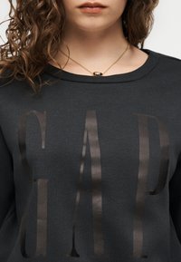 Ung person med krøllete hår iført en mørk genser med store, skinnende bokstaver "GAP" og et gyllent ovalt anhengsmykke.