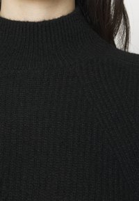 Gros plan sur un pull à col roulé noir côtelé porté par une personne aux longs cheveux foncés visibles sur le côté droit.