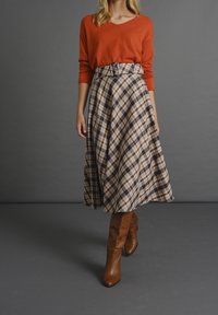 Pull couleur rouille avec col en V, assorti à une jupe plissée longueur genou aux tons beige, marron et noir. Bottes hautes marron jusqu'aux genoux.