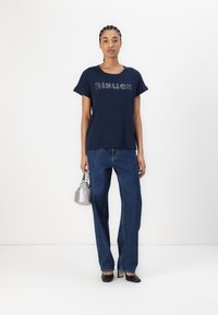 Mørkeblå bomull T-skjorte med korte ermer og "Blauer" trykket i sølv. Kombinert med høyhåndt blå jeans og holder en sølvfarget veske.