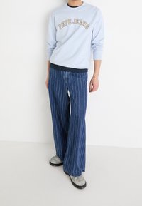 Sudadera azul claro con el logo "PEPE JEANS" y pantalones anchos de mezclilla con rayas azules. El calzado presenta un acabado texturizado de tono grisáceo.