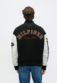 Άτομο με κατσαρά μαλλιά που φοράει μαύρο και άσπρο μπουφάν με την επιγραφή "HILFIGER Racing Club" στην πλάτη και μπλε τζιν, γυρισμένο με την πλάτη.