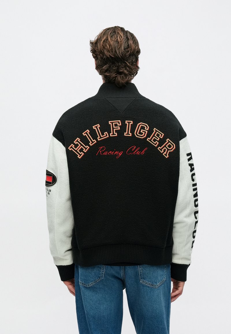 Άτομο με κατσαρά μαλλιά που φοράει μαύρο και άσπρο μπουφάν με την επιγραφή "HILFIGER Racing Club" στην πλάτη και μπλε τζιν, γυρισμένο με την πλάτη.